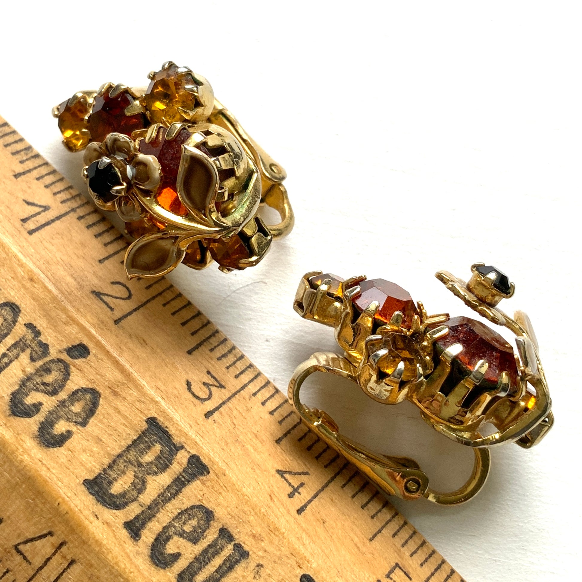 Clip-on earrings featuring vibrant orange and amber rhinestones, adorned with a petite beige enamel flower.  小さなベージュのエナメルフラワーが咲いた、アンバーラインストーンのクリップイヤリング。