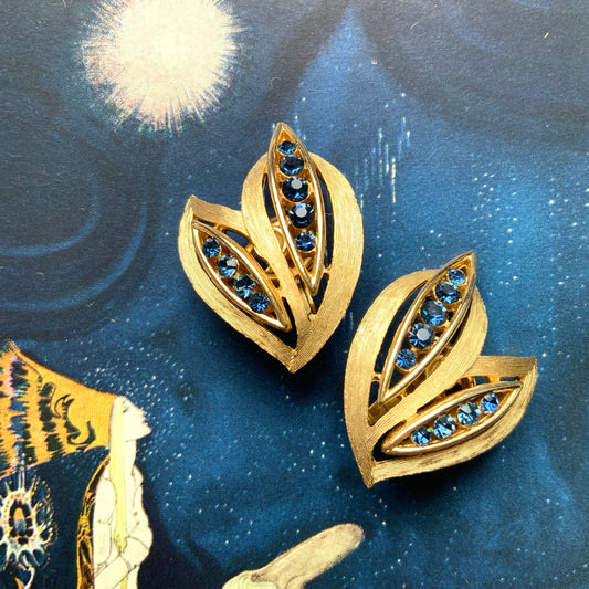 Trifari clip-on earrings featuring slim, bamboo leaf–like gold-tone leaves adorned with a row of blue rhinestones. 笹の葉のようにスリムなゴールドトーンのリーフに、ブルーのラインストーンが並んだトリファリのイヤリング。 