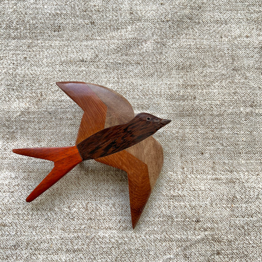 A wooden swallow brooch made from different shades of natural wood. 色味の異なる木材を組み合わせて作られた、ツバメモチーフのウッドブローチ。