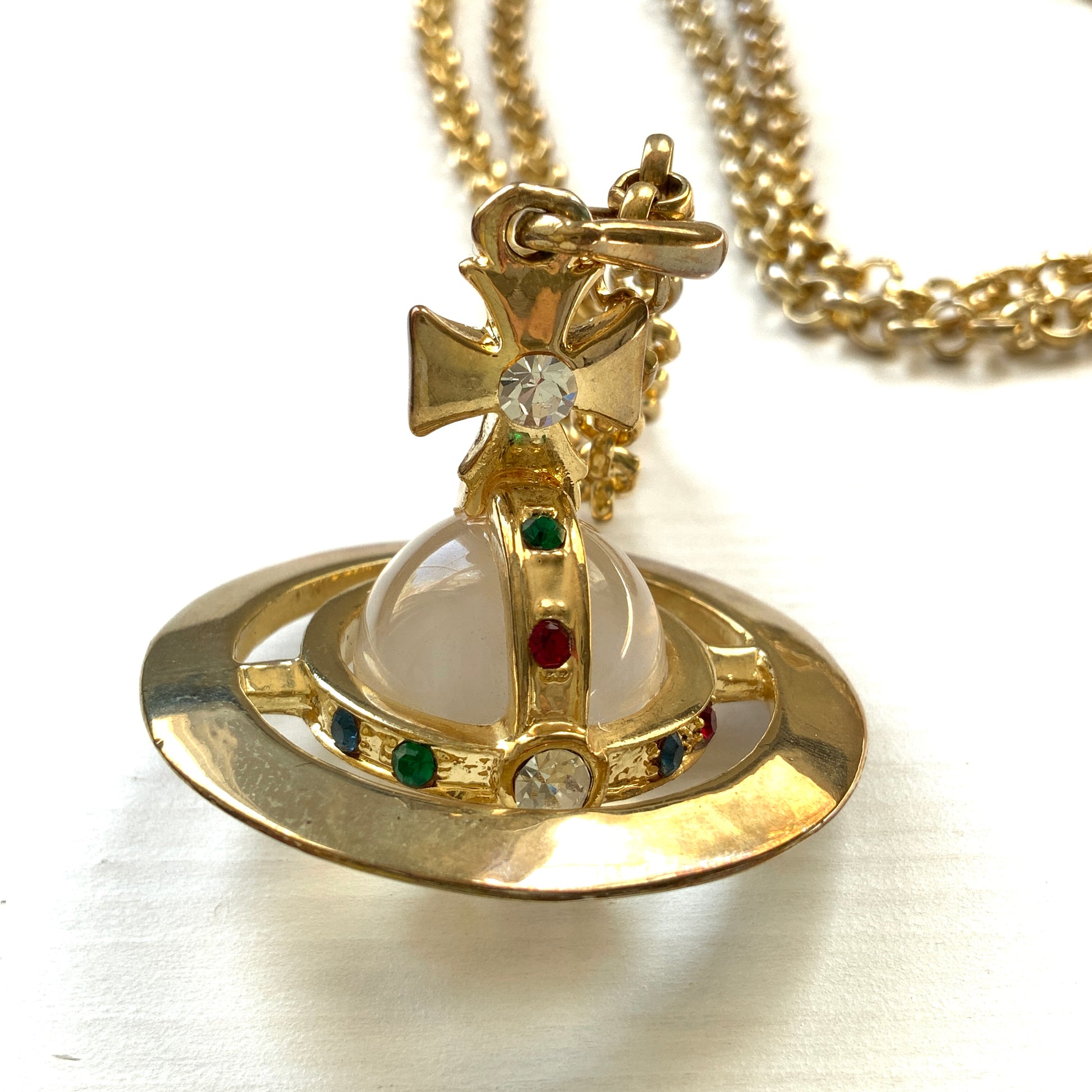A striking pendant necklace featuring a decorative orb encircled by a metal hoop, resembling the rings of Saturn. カラフルなストーンで飾られた宝珠（キリスト教圏で、十字架をのせた球体として王権の象徴に用いられるモチーフ）を、まるで土星の輪のようなメタルフープで囲んだデザインのペンダントネックレスです。