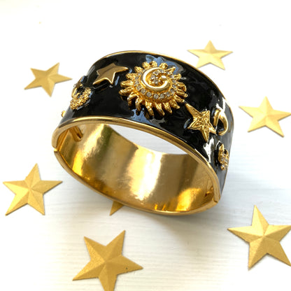A wide bangle featuring celestial motifs in gold tone set against glossy black enamel. ブラックエナメルに、ゴールドトーンの天体モチーフが輝く幅広バングル。 
