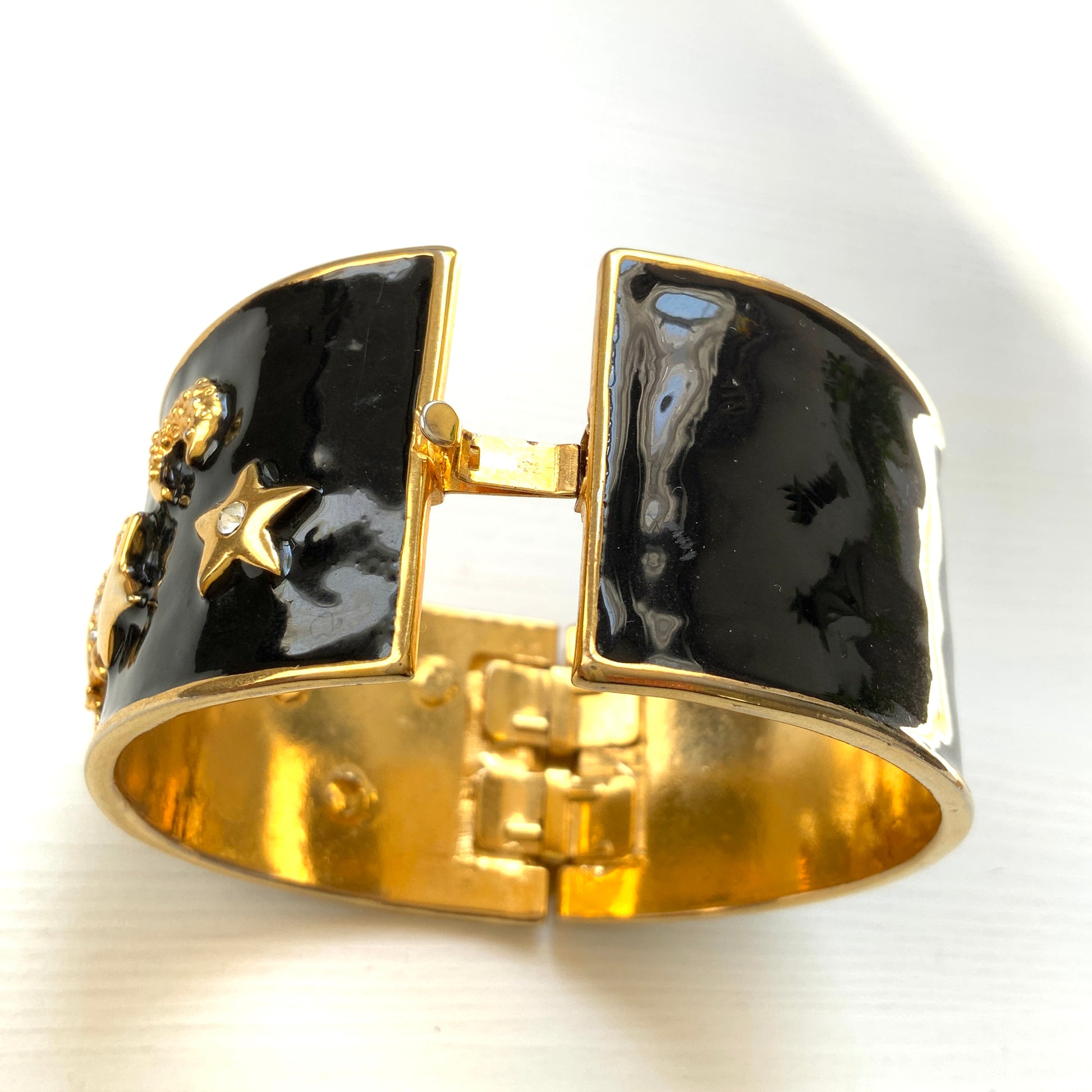 A wide bangle featuring celestial motifs in gold tone set against glossy black enamel. ブラックエナメルに、ゴールドトーンの天体モチーフが輝く幅広バングル。 