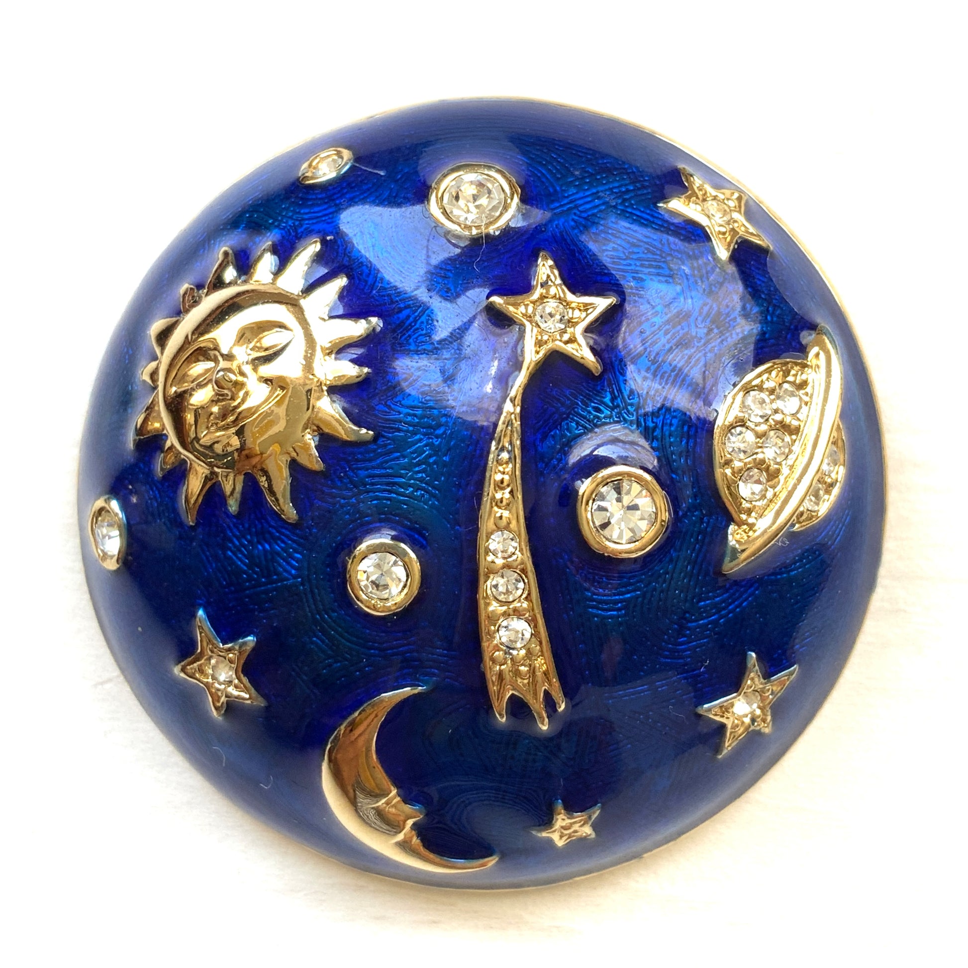 A dome-shaped brooch adorned with vibrant blue enamel, featuring silver-tone celestial motifs such as the sun face, crescent moon face, Saturn, and stars. 鮮やかなブルーのエナメルが施されたドームに、シルバートーンの太陽や三日月フェイスや、土星や星などが浮かんだブローチ。