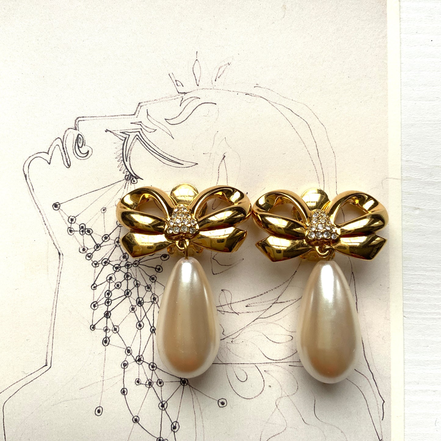 A pair of clip-on earrings featuring a bow motif in gold-tone metal, with large teardrop-shaped faux pearls that sway beautifully as you move. ゴールドトーンメタルのリボンモチーフに、大ぶりのティアドロップ型フォーパールが揺れるクリップイヤリング。 