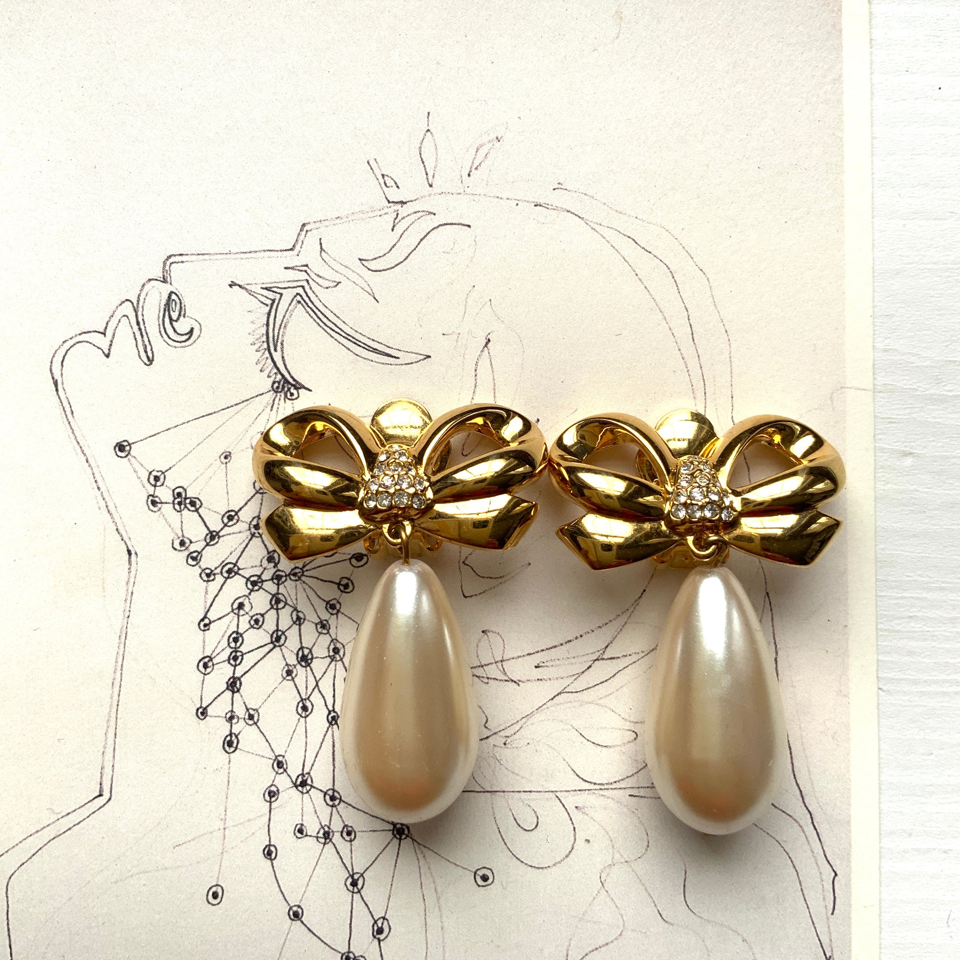 A pair of clip-on earrings featuring a bow motif in gold-tone metal, with large teardrop-shaped faux pearls that sway beautifully as you move. ゴールドトーンメタルのリボンモチーフに、大ぶりのティアドロップ型フォーパールが揺れるクリップイヤリング。 