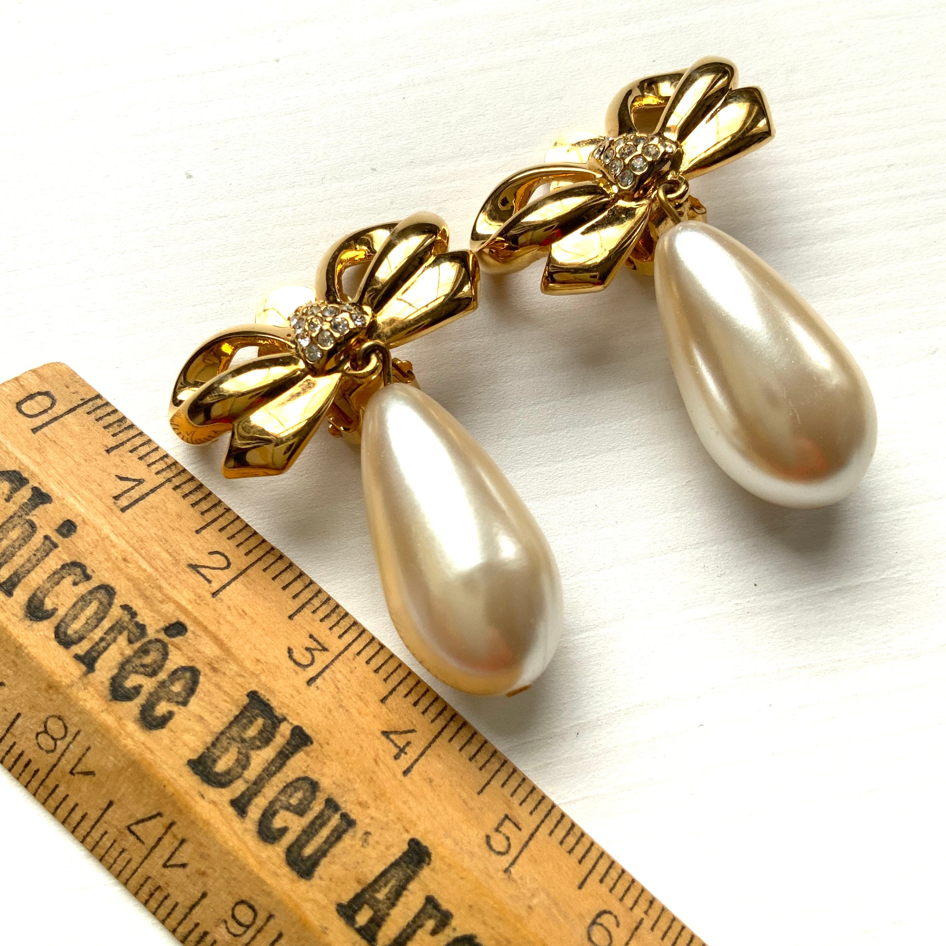 A pair of clip-on earrings featuring a bow motif in gold-tone metal, with large teardrop-shaped faux pearls that sway beautifully as you move. ゴールドトーンメタルのリボンモチーフに、大ぶりのティアドロップ型フォーパールが揺れるクリップイヤリング。 