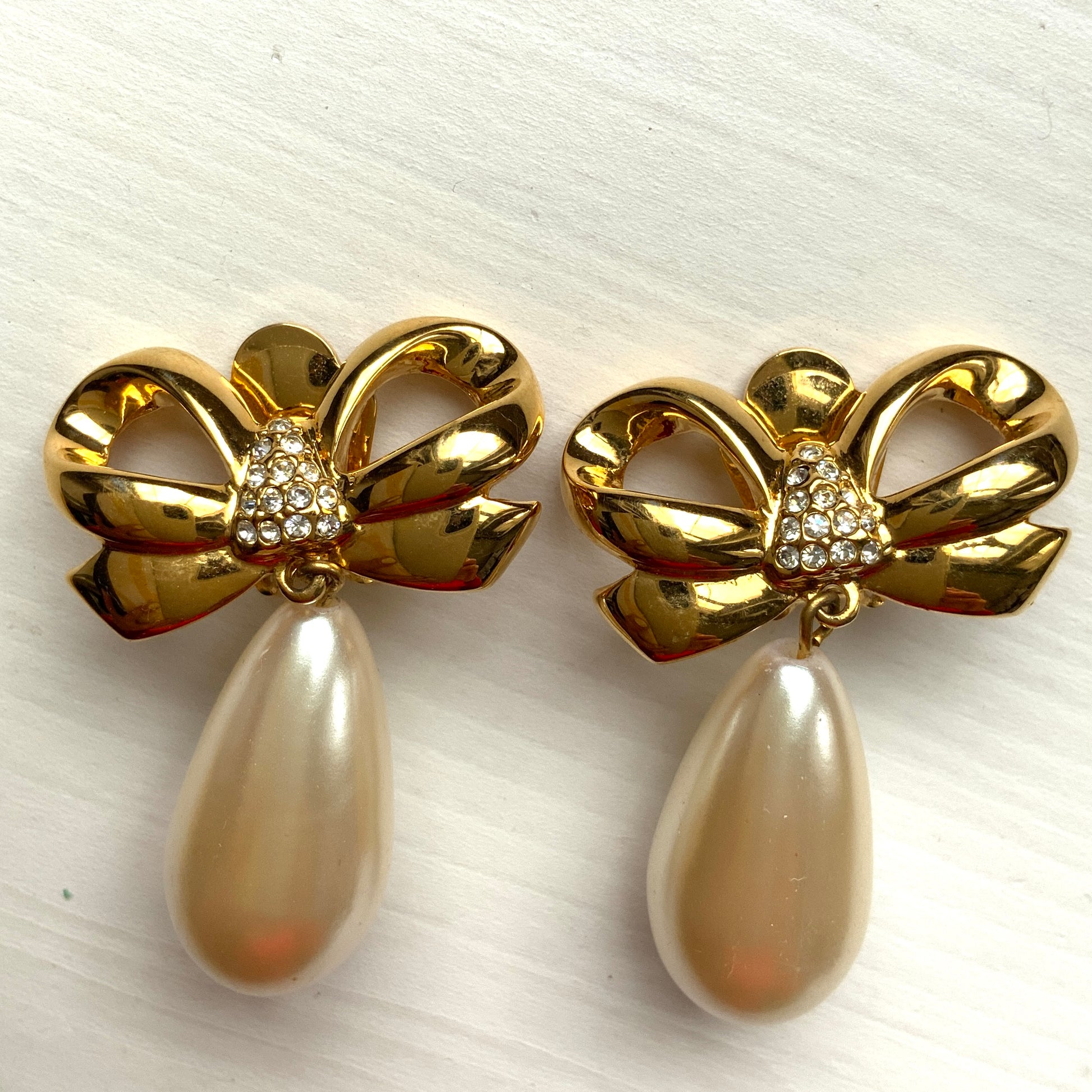 A pair of clip-on earrings featuring a bow motif in gold-tone metal, with large teardrop-shaped faux pearls that sway beautifully as you move. ゴールドトーンメタルのリボンモチーフに、大ぶりのティアドロップ型フォーパールが揺れるクリップイヤリング。 