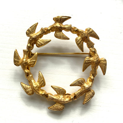A gold-tone wreath brooch formed by a circle of small birds in flight. ゴールドトーンメタルでつくられた、鳥たちのリースブローチ。 