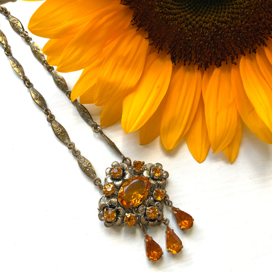 An oval amber glass jewel is surrounded by delicate metal flowers, with matching amber teardrop rhinestones gently dangling below. オーバル型のアンバーグラスビジューの周りをメタルのお花が取り囲み、同じアンバーカラーのティアドロップラインストーンが揺れるネックレス。 