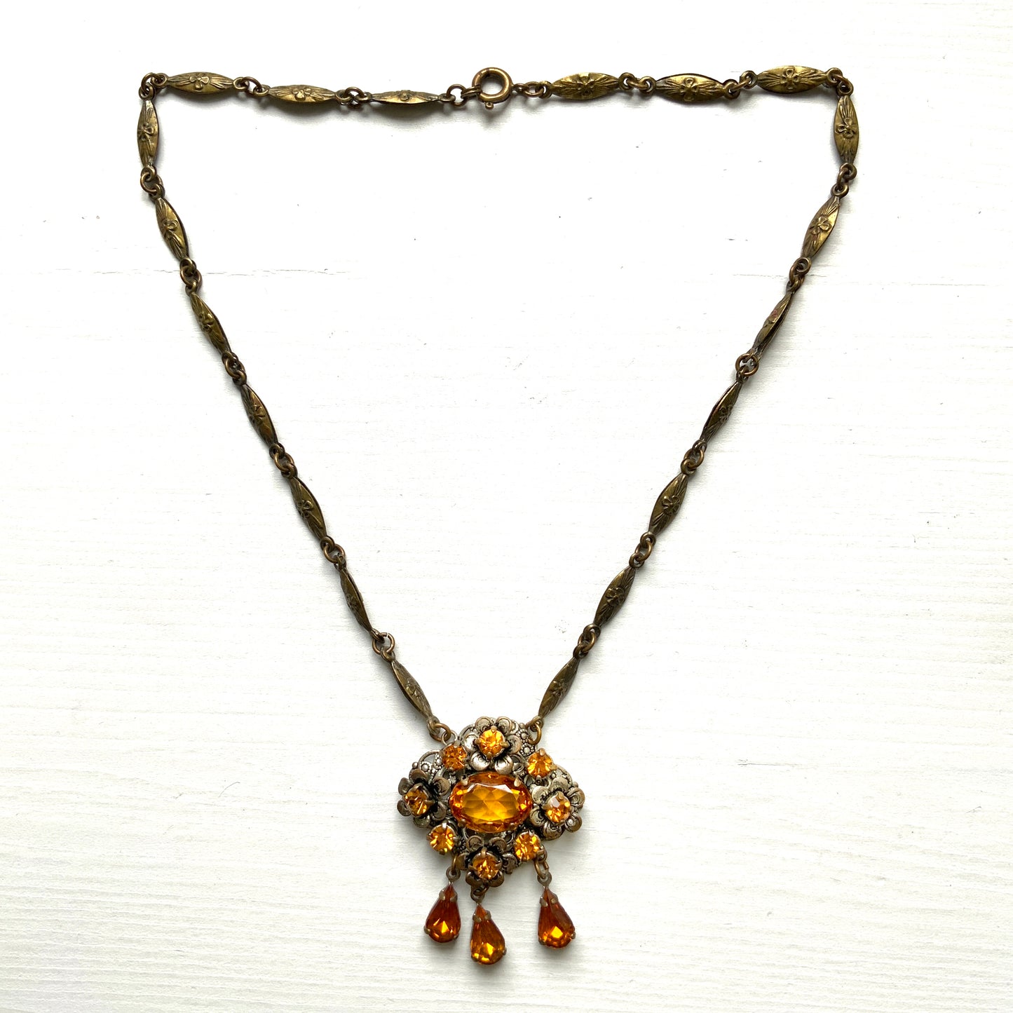 An oval amber glass jewel is surrounded by delicate metal flowers, with matching amber teardrop rhinestones gently dangling below. オーバル型のアンバーグラスビジューの周りをメタルのお花が取り囲み、同じアンバーカラーのティアドロップラインストーンが揺れるネックレス。 