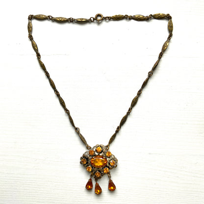 An oval amber glass jewel is surrounded by delicate metal flowers, with matching amber teardrop rhinestones gently dangling below. オーバル型のアンバーグラスビジューの周りをメタルのお花が取り囲み、同じアンバーカラーのティアドロップラインストーンが揺れるネックレス。 
