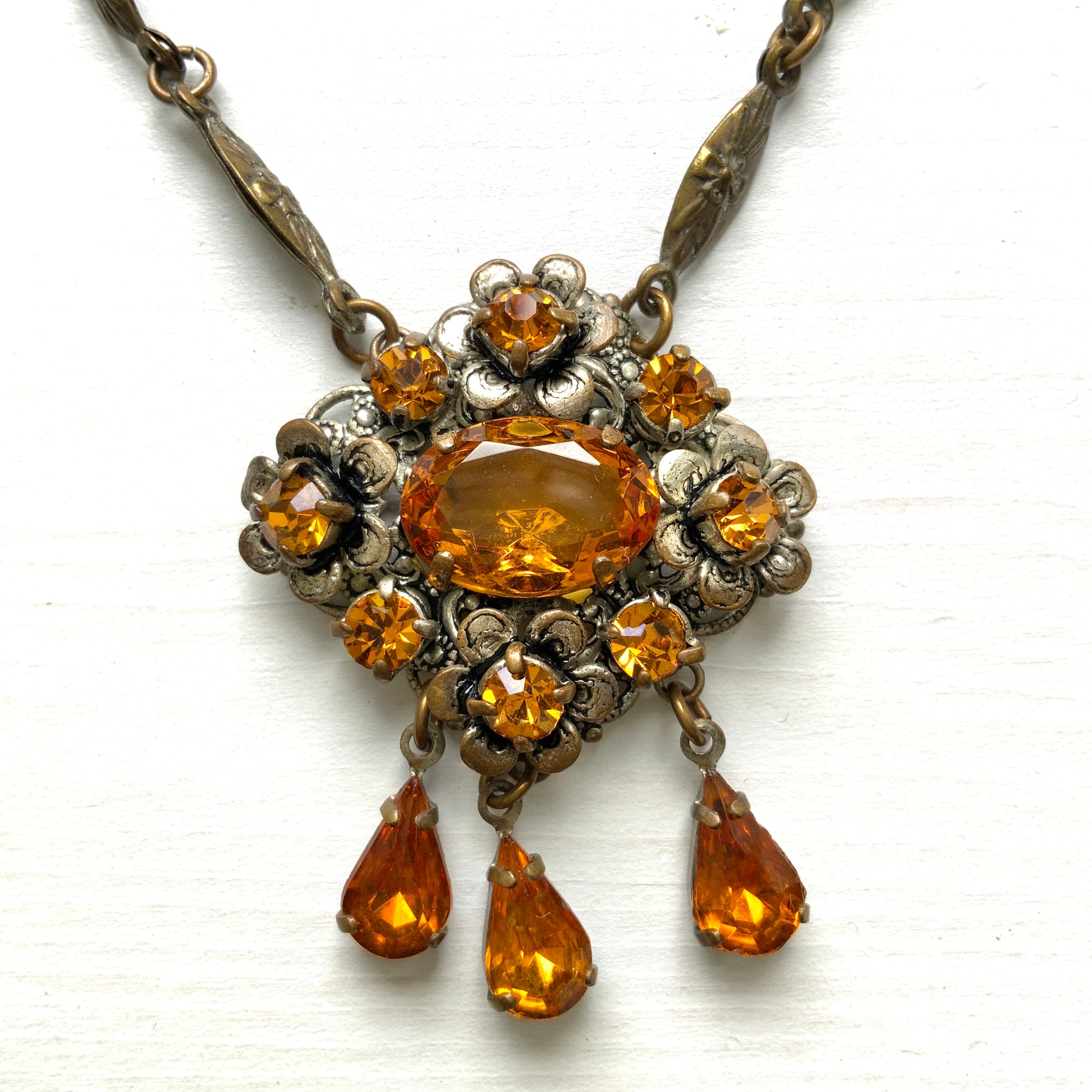 An oval amber glass jewel is surrounded by delicate metal flowers, with matching amber teardrop rhinestones gently dangling below. オーバル型のアンバーグラスビジューの周りをメタルのお花が取り囲み、同じアンバーカラーのティアドロップラインストーンが揺れるネックレス。 