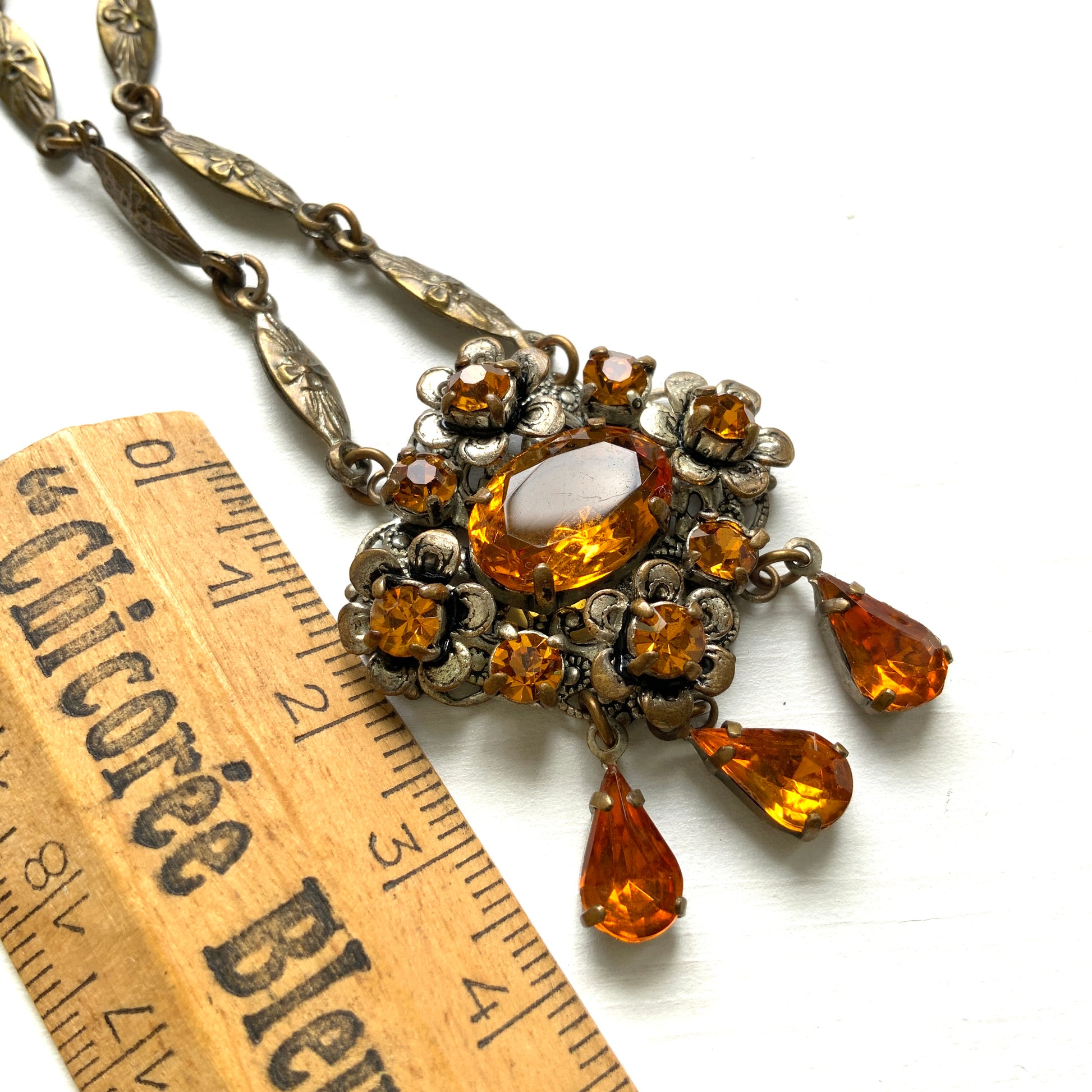 An oval amber glass jewel is surrounded by delicate metal flowers, with matching amber teardrop rhinestones gently dangling below. オーバル型のアンバーグラスビジューの周りをメタルのお花が取り囲み、同じアンバーカラーのティアドロップラインストーンが揺れるネックレス。 