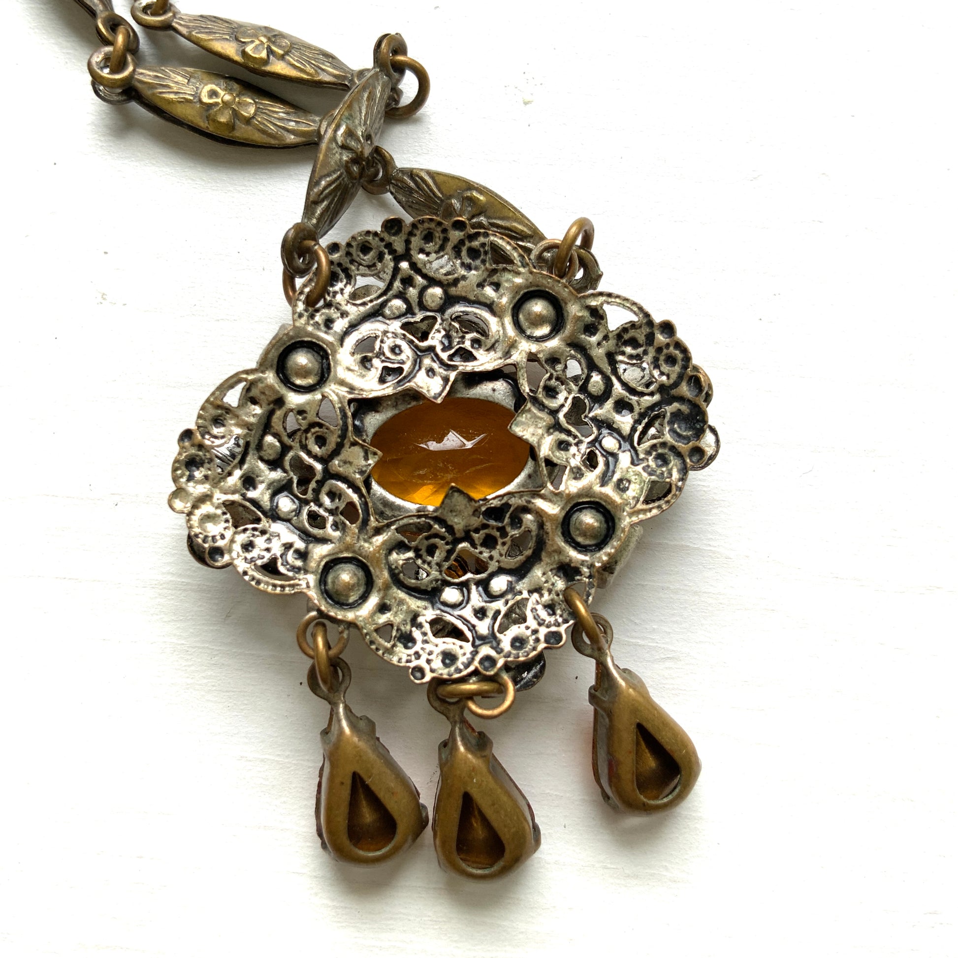An oval amber glass jewel is surrounded by delicate metal flowers, with matching amber teardrop rhinestones gently dangling below. オーバル型のアンバーグラスビジューの周りをメタルのお花が取り囲み、同じアンバーカラーのティアドロップラインストーンが揺れるネックレス。 