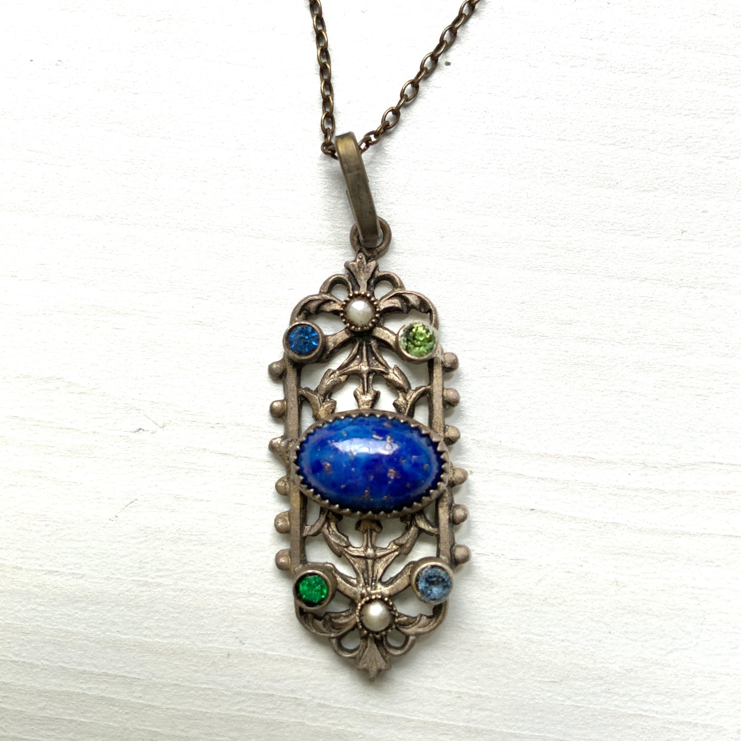 A delicate filigree pendant necklace adorned with a lapis glass cabochon, colourful rhinestones, and seed glass pearls. 繊細なフィリグリーのプレートに、ラピスグラスのカボションとカラフルなラインストーン、シードグラスパールがあしらわれたペンダントネックレス。 