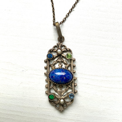 A delicate filigree pendant necklace adorned with a lapis glass cabochon, colourful rhinestones, and seed glass pearls. 繊細なフィリグリーのプレートに、ラピスグラスのカボションとカラフルなラインストーン、シードグラスパールがあしらわれたペンダントネックレス。 