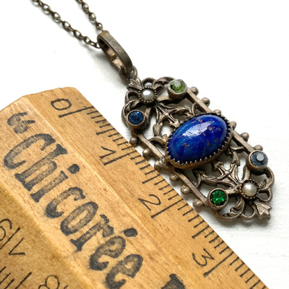 A delicate filigree pendant necklace adorned with a lapis glass cabochon, colourful rhinestones, and seed glass pearls. 繊細なフィリグリーのプレートに、ラピスグラスのカボションとカラフルなラインストーン、シードグラスパールがあしらわれたペンダントネックレス。 
