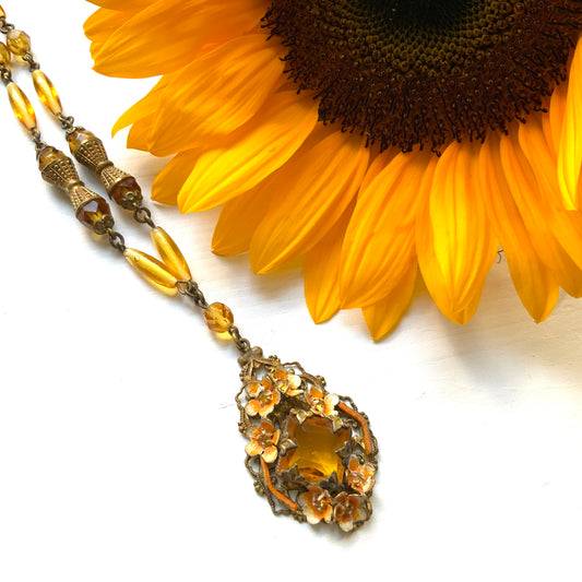 A Czech-made necklace featuring a diamond-shaped amber glass stone encircled by vivid orange enamel flowers. ダイヤ型のアンバーグラスをオレンジ色のエナメルフラワーが取り囲むチェコ製のネックレス。 