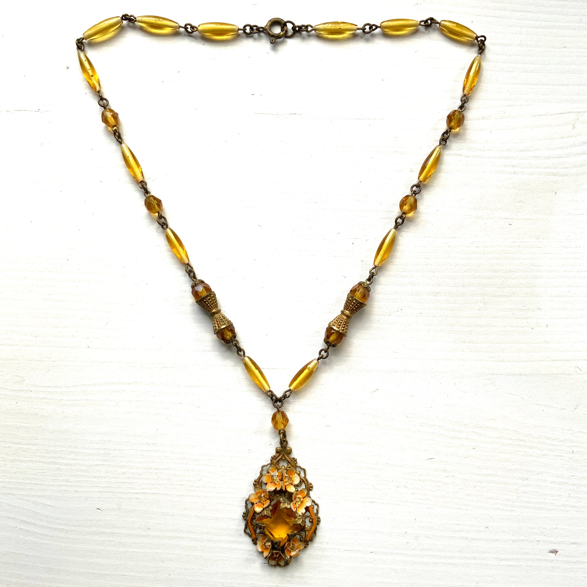 A Czech-made necklace featuring a diamond-shaped amber glass stone encircled by vivid orange enamel flowers. ダイヤ型のアンバーグラスをオレンジ色のエナメルフラワーが取り囲むチェコ製のネックレス。 