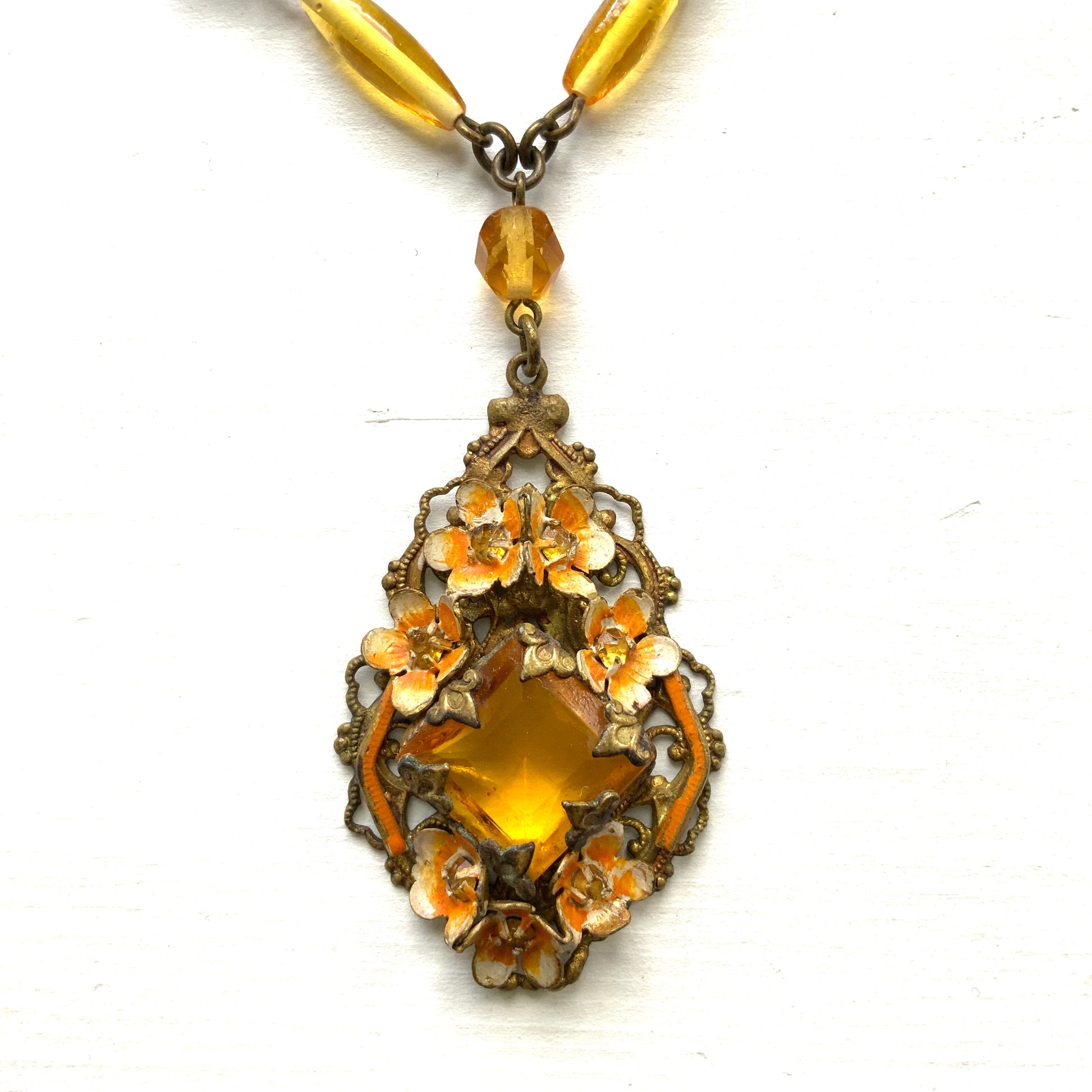 A Czech-made necklace featuring a diamond-shaped amber glass stone encircled by vivid orange enamel flowers. ダイヤ型のアンバーグラスをオレンジ色のエナメルフラワーが取り囲むチェコ製のネックレス。 