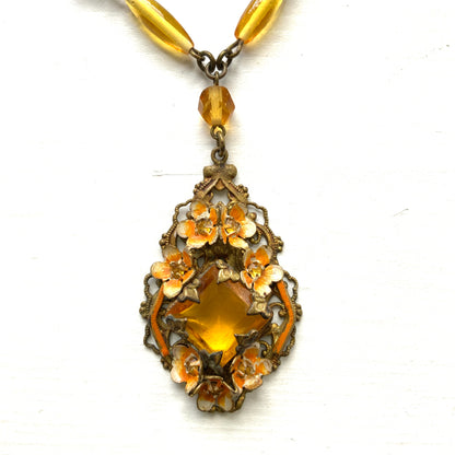 A Czech-made necklace featuring a diamond-shaped amber glass stone encircled by vivid orange enamel flowers. ダイヤ型のアンバーグラスをオレンジ色のエナメルフラワーが取り囲むチェコ製のネックレス。 