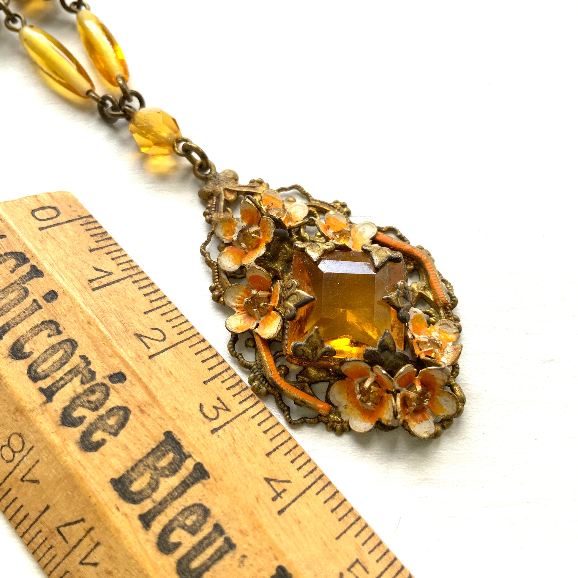 A Czech-made necklace featuring a diamond-shaped amber glass stone encircled by vivid orange enamel flowers. ダイヤ型のアンバーグラスをオレンジ色のエナメルフラワーが取り囲むチェコ製のネックレス。 