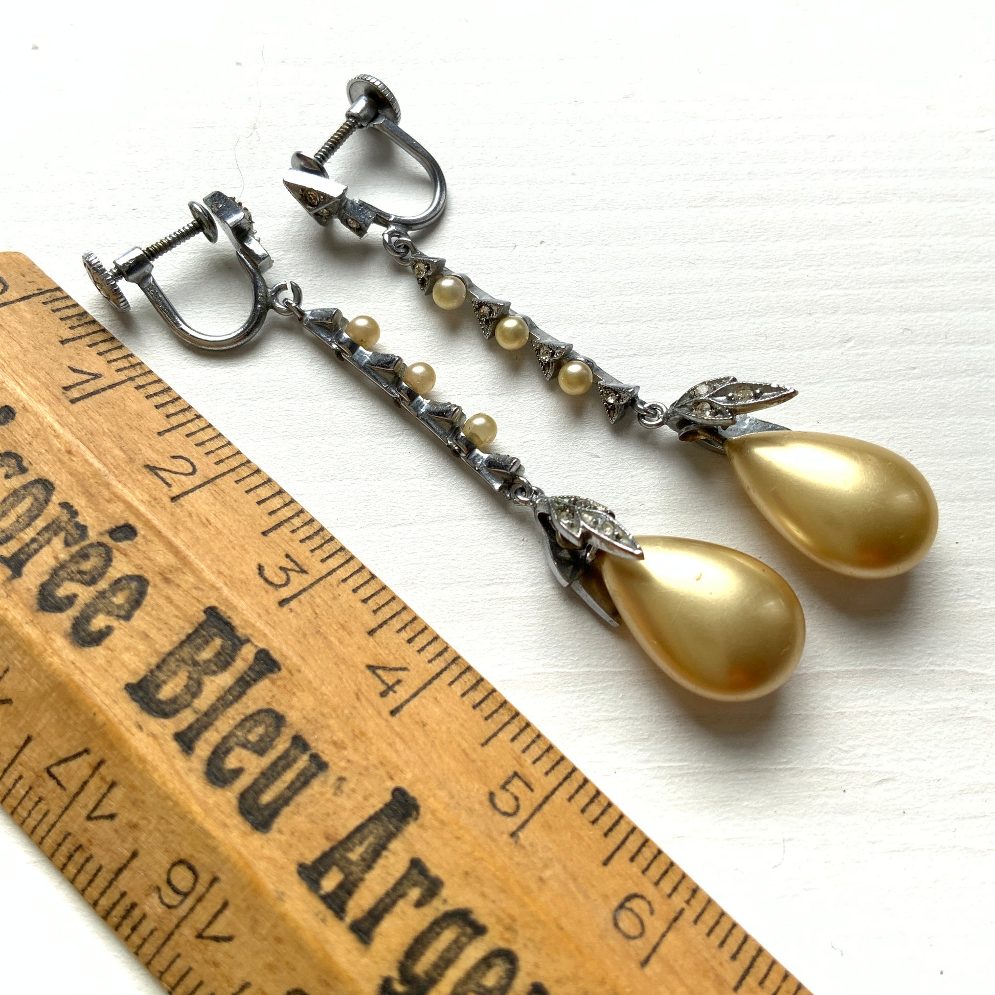 Screw earrings featuring a silver-tone leaf with a champagne gold teardrop pearl glass dangling from the tip. シルバートーンのリーフの先にシャンパンゴールドカラーのティアドロップグラスパールが揺れるスクリューイヤリング。
