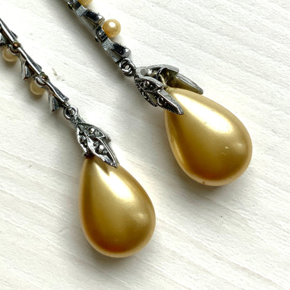 Screw earrings featuring a silver-tone leaf with a champagne gold teardrop pearl glass dangling from the tip. シルバートーンのリーフの先にシャンパンゴールドカラーのティアドロップグラスパールが揺れるスクリューイヤリング。