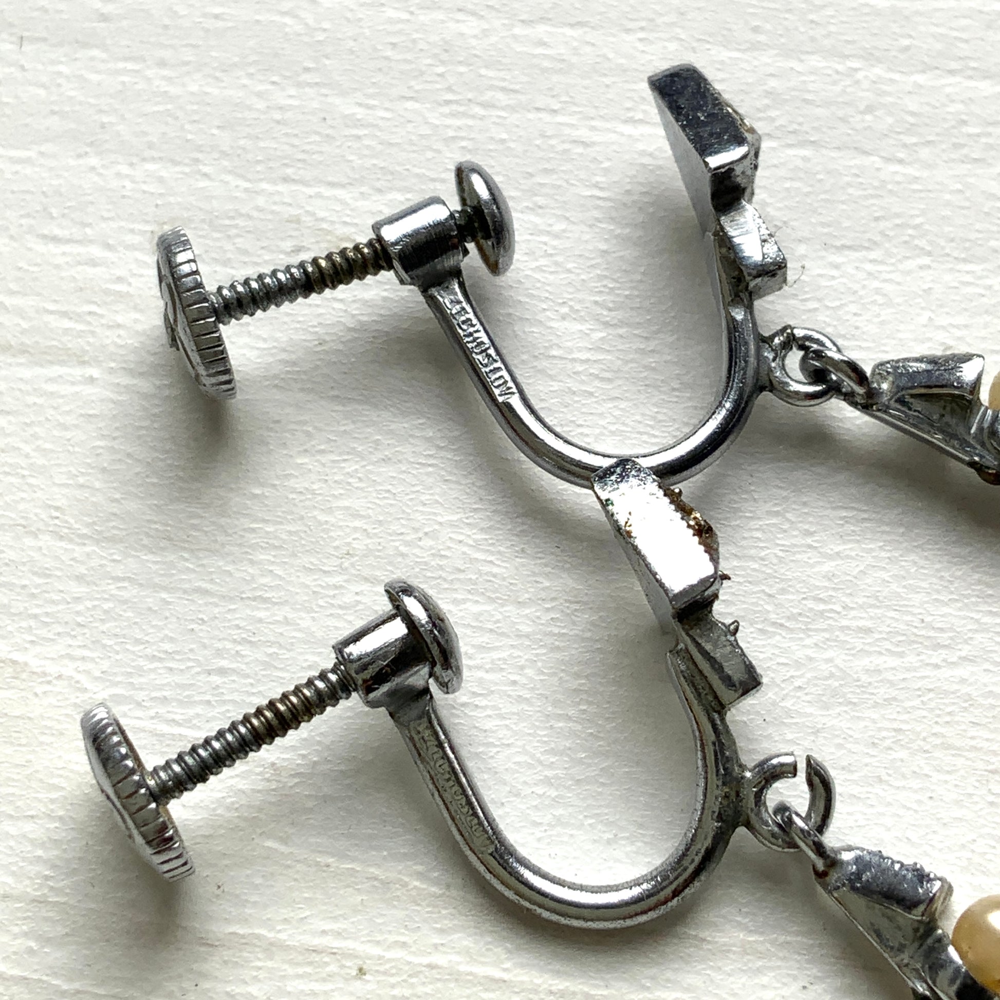 Screw earrings featuring a silver-tone leaf with a champagne gold teardrop pearl glass dangling from the tip. シルバートーンのリーフの先にシャンパンゴールドカラーのティアドロップグラスパールが揺れるスクリューイヤリング。