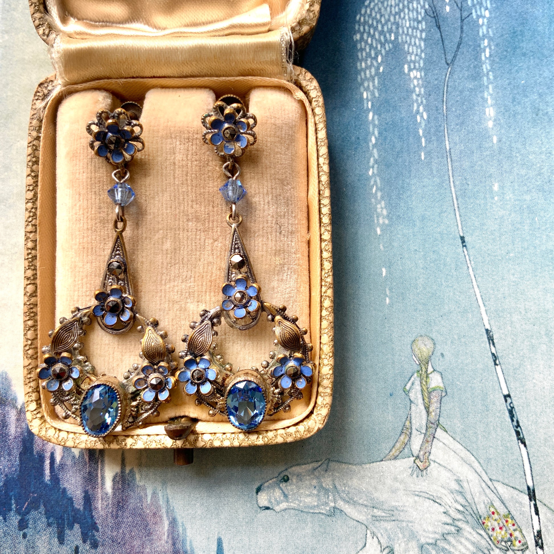 Screw-back earrings featuring pale blue enamel flowers and glass bijoux. Delicately crafted in a crescent-shaped filigree, the tiny enamel blossoms sparkle alongside drops of soft, water-like blue glass stones. 淡いブルーのエナメルフラワーとグラスビジューのスクリューイヤリング。 三日月型の繊細なフィリグリーに、小さなエナメルの花々と、水の雫のように澄んだブルーグラスがきらめきます。 