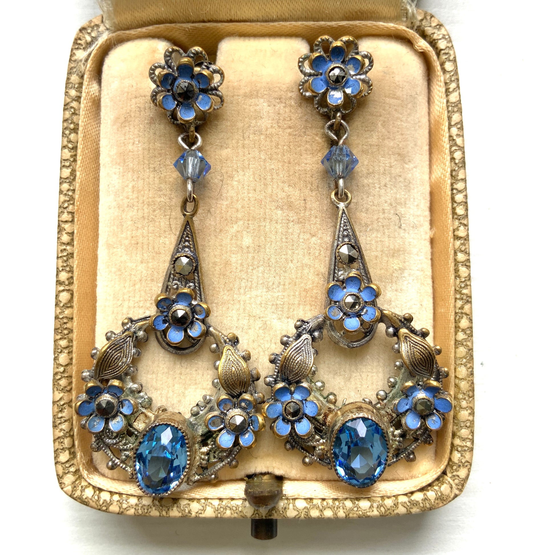 Screw-back earrings featuring pale blue enamel flowers and glass bijoux. Delicately crafted in a crescent-shaped filigree, the tiny enamel blossoms sparkle alongside drops of soft, water-like blue glass stones. 淡いブルーのエナメルフラワーとグラスビジューのスクリューイヤリング。 三日月型の繊細なフィリグリーに、小さなエナメルの花々と、水の雫のように澄んだブルーグラスがきらめきます。 