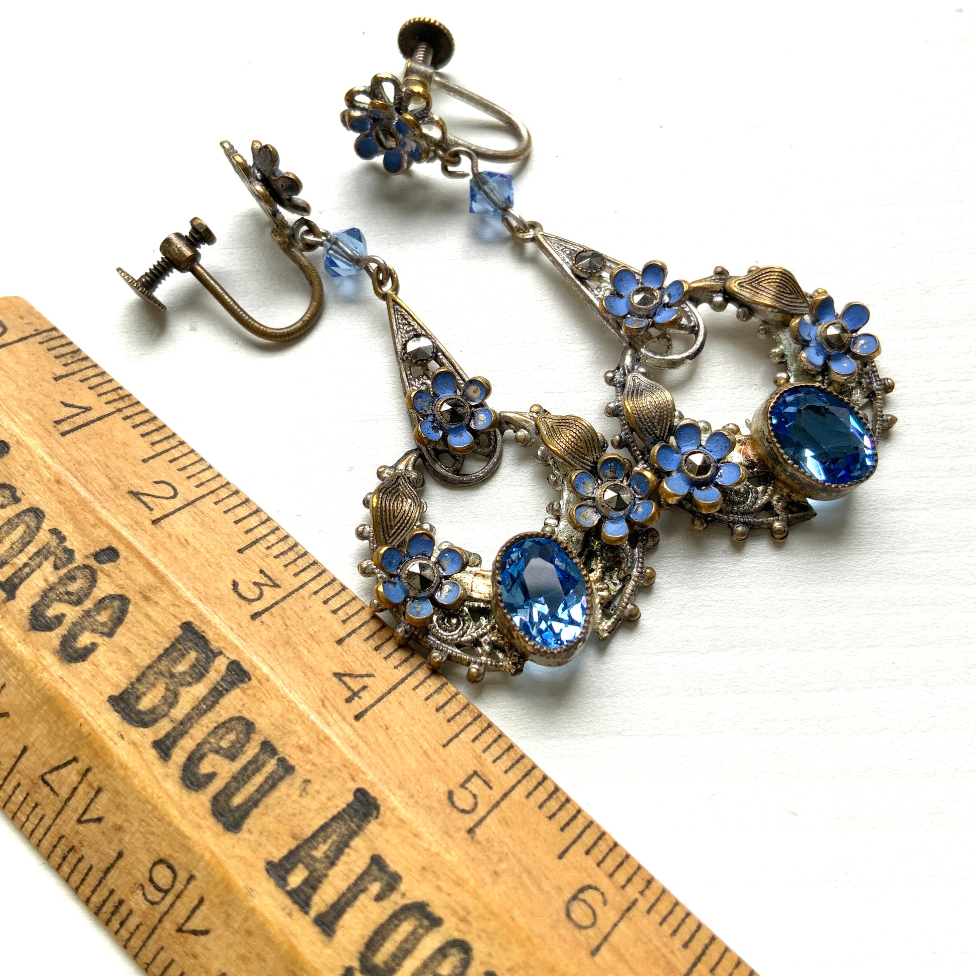 Screw-back earrings featuring pale blue enamel flowers and glass bijoux. Delicately crafted in a crescent-shaped filigree, the tiny enamel blossoms sparkle alongside drops of soft, water-like blue glass stones. 淡いブルーのエナメルフラワーとグラスビジューのスクリューイヤリング。 三日月型の繊細なフィリグリーに、小さなエナメルの花々と、水の雫のように澄んだブルーグラスがきらめきます。 
