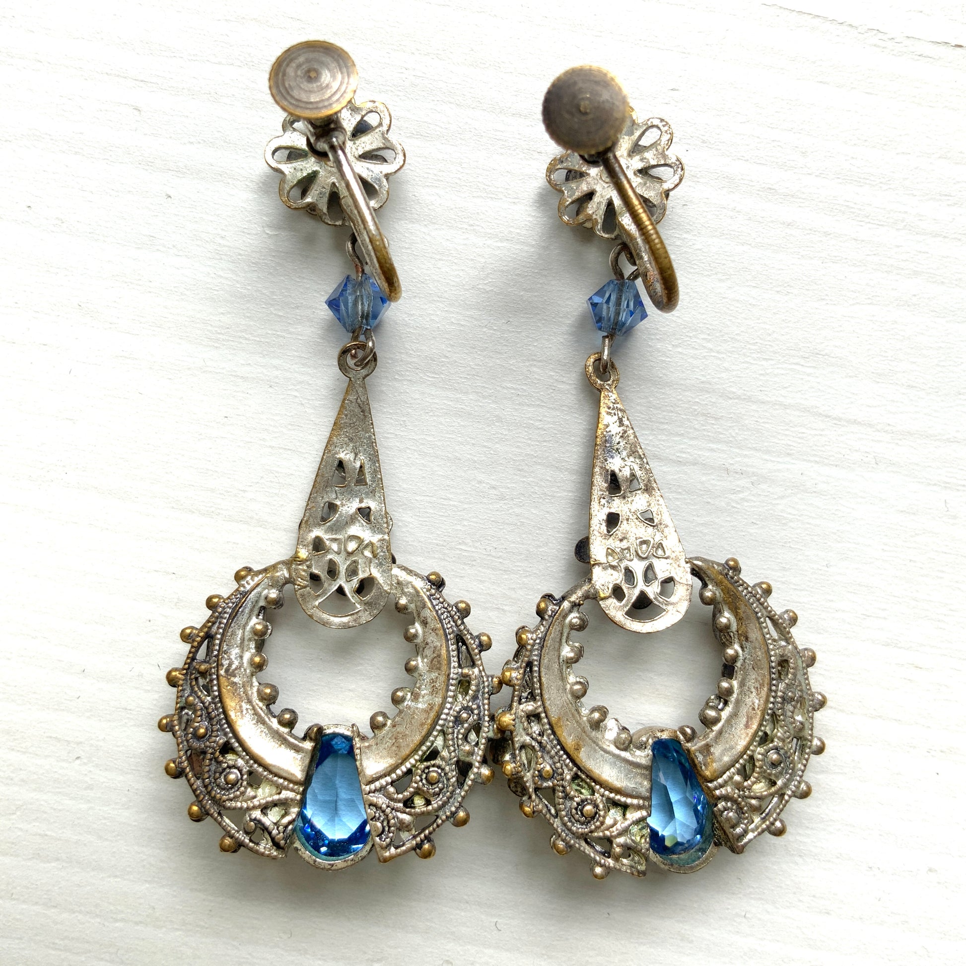 Screw-back earrings featuring pale blue enamel flowers and glass bijoux. Delicately crafted in a crescent-shaped filigree, the tiny enamel blossoms sparkle alongside drops of soft, water-like blue glass stones. 淡いブルーのエナメルフラワーとグラスビジューのスクリューイヤリング。 三日月型の繊細なフィリグリーに、小さなエナメルの花々と、水の雫のように澄んだブルーグラスがきらめきます。 