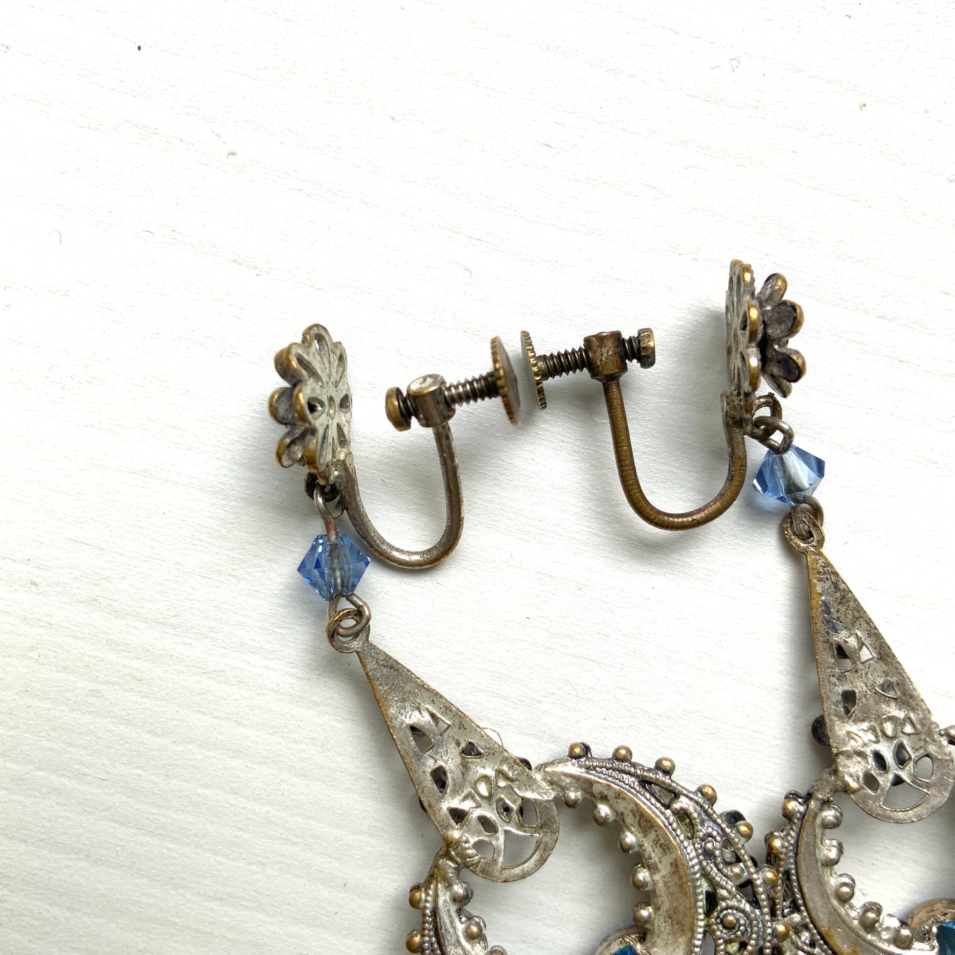 Screw-back earrings featuring pale blue enamel flowers and glass bijoux. Delicately crafted in a crescent-shaped filigree, the tiny enamel blossoms sparkle alongside drops of soft, water-like blue glass stones. 淡いブルーのエナメルフラワーとグラスビジューのスクリューイヤリング。 三日月型の繊細なフィリグリーに、小さなエナメルの花々と、水の雫のように澄んだブルーグラスがきらめきます。 