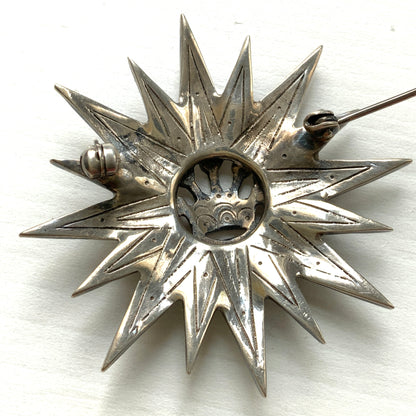 A silver brooch featuring a small crown at the centre of a starburst design. スターバーストの中央に小さな王冠があしらわれた、シルバー製のブローチ。