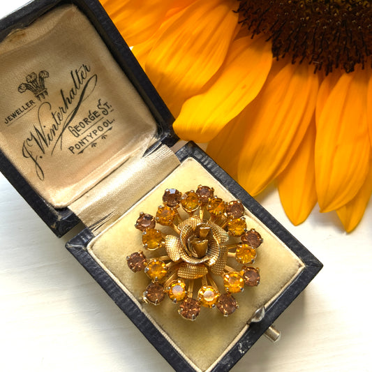 A vintage brooch featuring a small gold-tone rose, with two-tone amber rhinestones sparkling out like a burst of light. ゴールドトーンの小さなバラから２トーンのアンバーラインストーンがスパークしているようなブローチ。