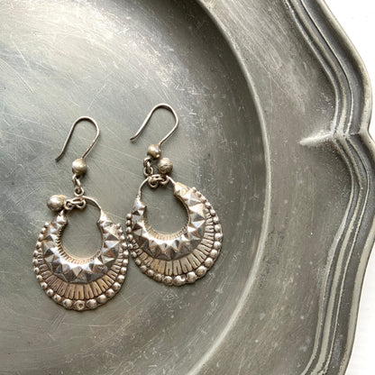 Striking silver tone hoop earrings with geometric embossed patterns and granulated bead details. 半月型にジオメトリックな打ち出し模様と粒状の装飾を施した、存在感あるシルバートーンのフープピアス。