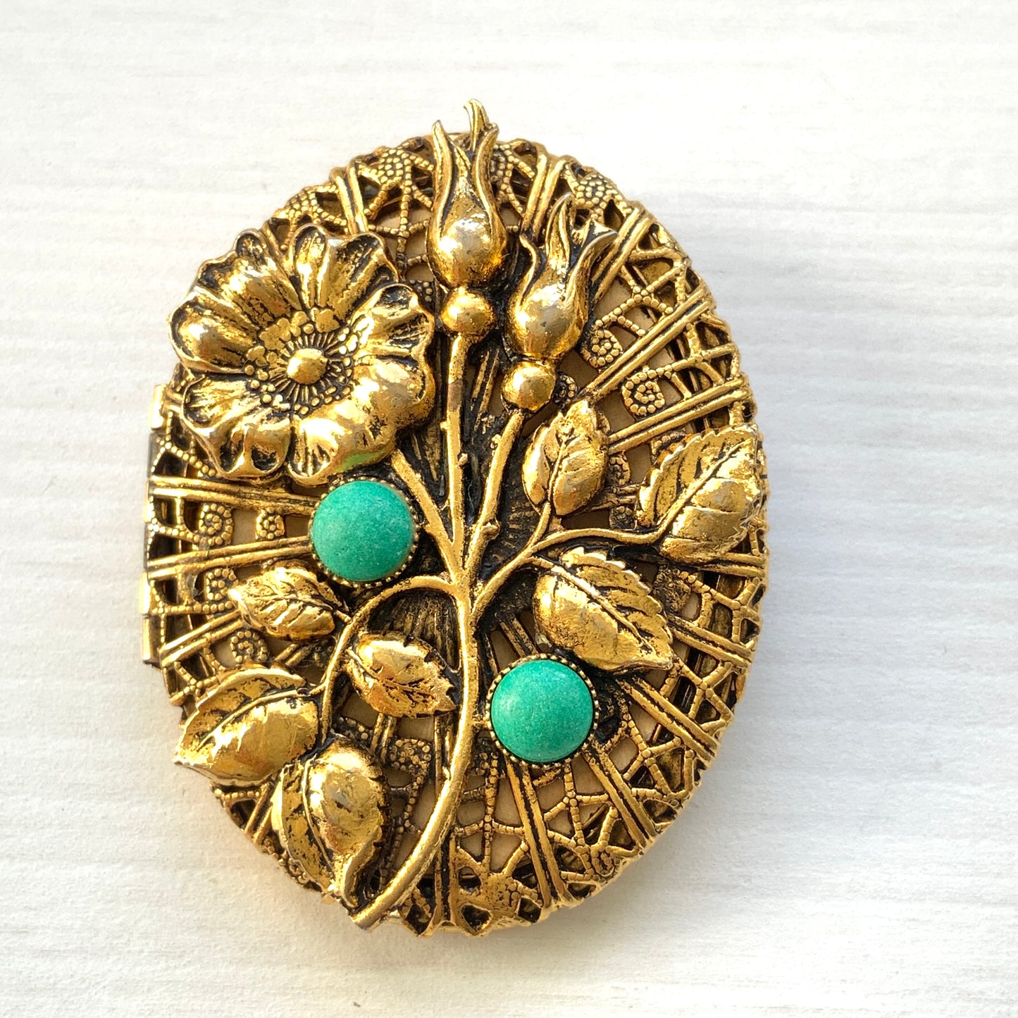 A gold-tone locket brooch featuring a floral motif accented with turquoise blue cabochons.  ゴールドトーンのお花に、ターコイズブルーのカボションが映えるロケットブローチ。