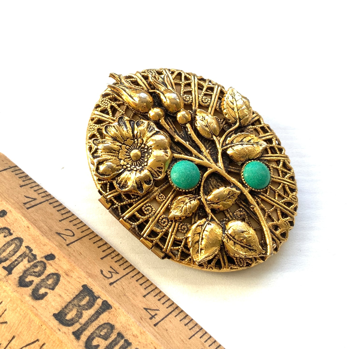A gold-tone locket brooch featuring a floral motif accented with turquoise blue cabochons.  ゴールドトーンのお花に、ターコイズブルーのカボションが映えるロケットブローチ。
